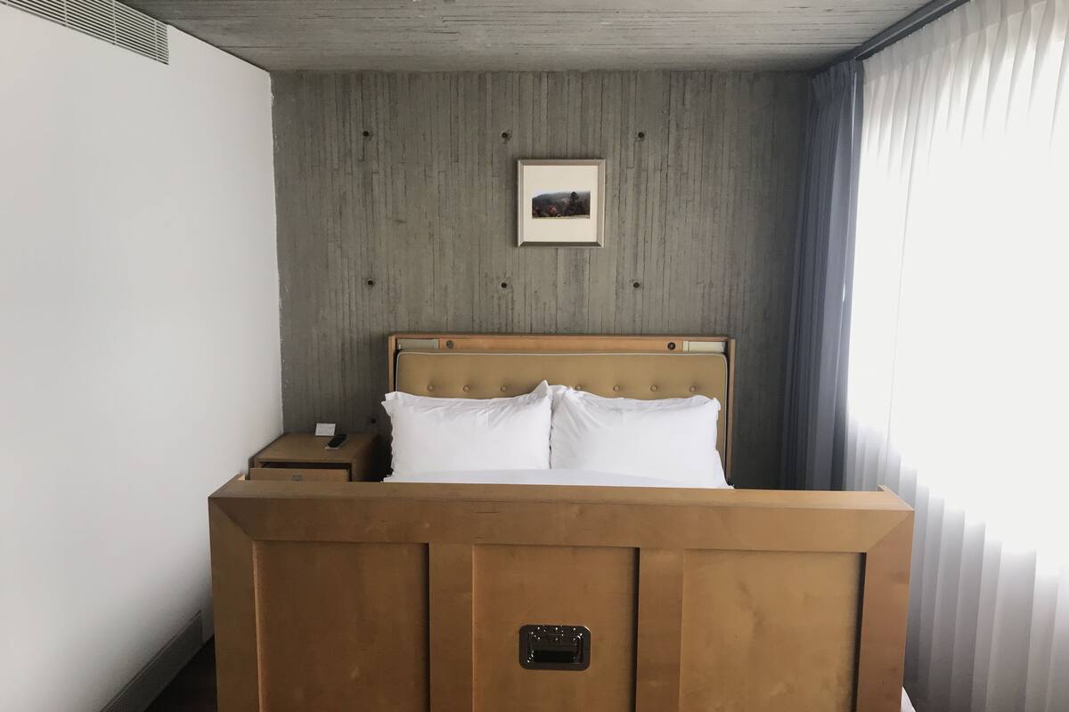 Una habitación de hotel con una cama de madera, una mesita de noche pequeña, una lámpara de noche, un cuadro en la pared y una ventana con cortinas.