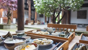 En traditionel japansk måltidsopstilling med flere retter og redskaber på et træbord, placeret i en fredfyldt udendørs have med træer og en bygning i baggrunden.