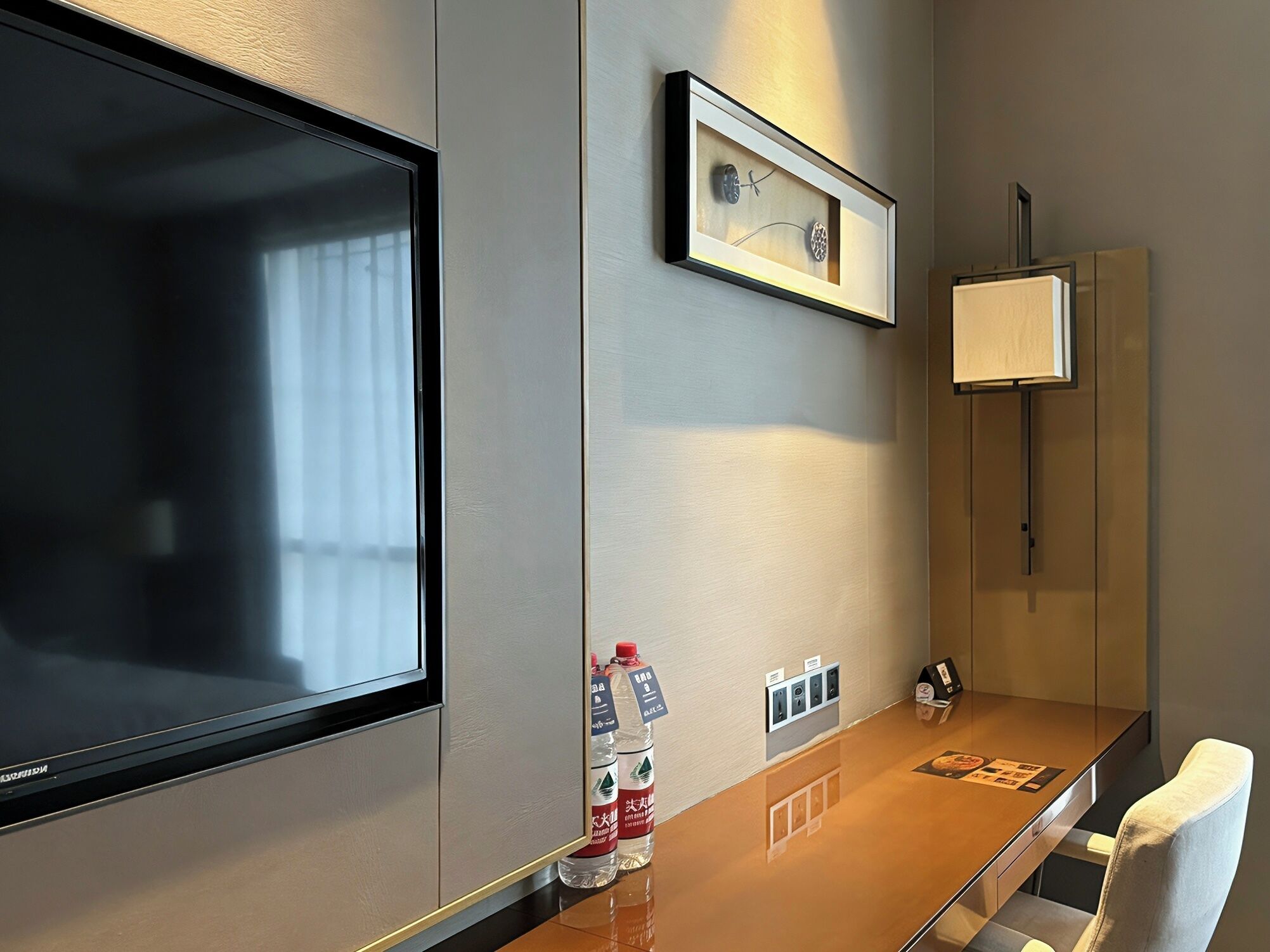 A hotel room with a large flat-screen TV, a framed picture on the wall, a minibar, a water dispenser, and a wooden table with a chair.