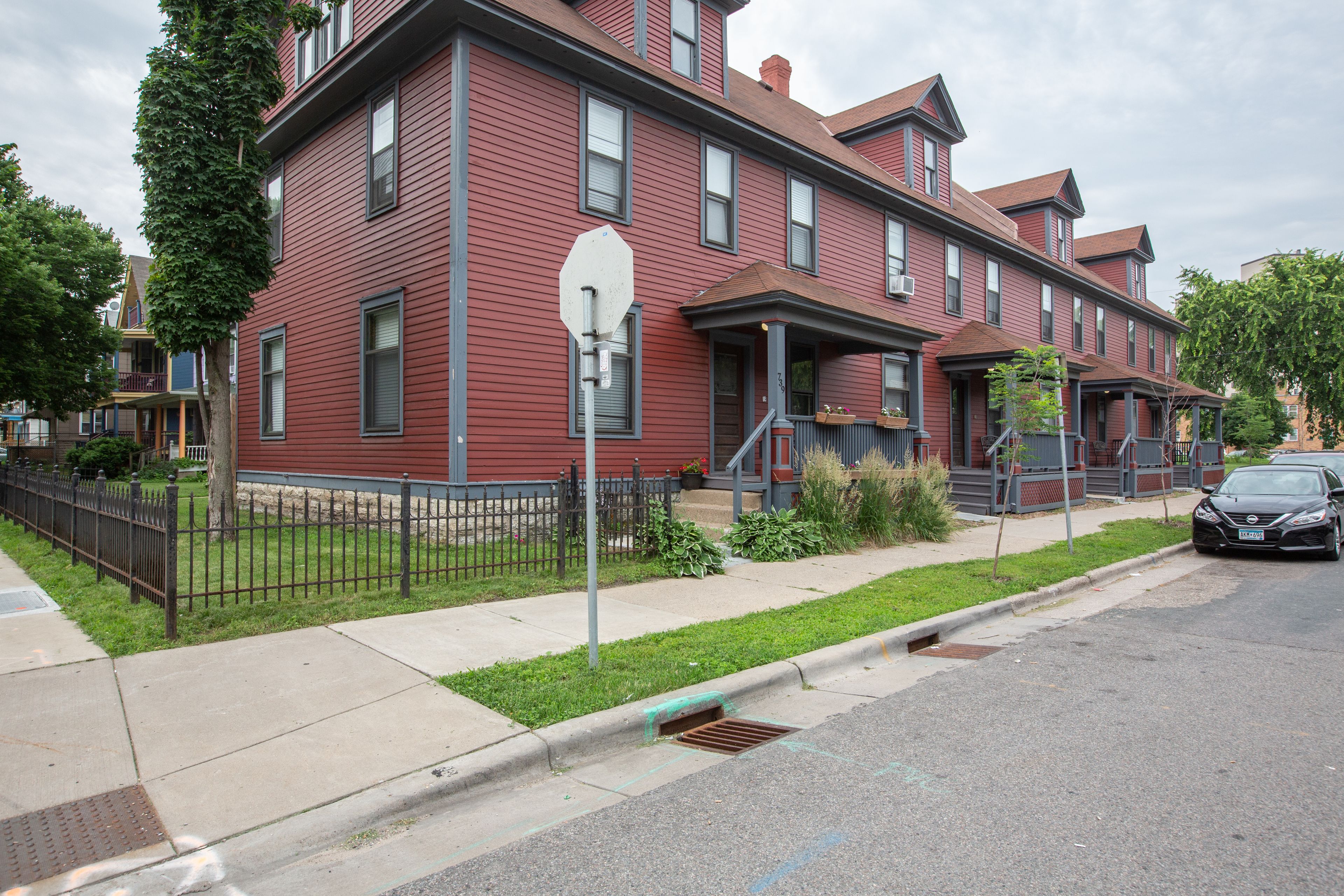 A red two-story building with a front porch, a small garden, and a parked car.