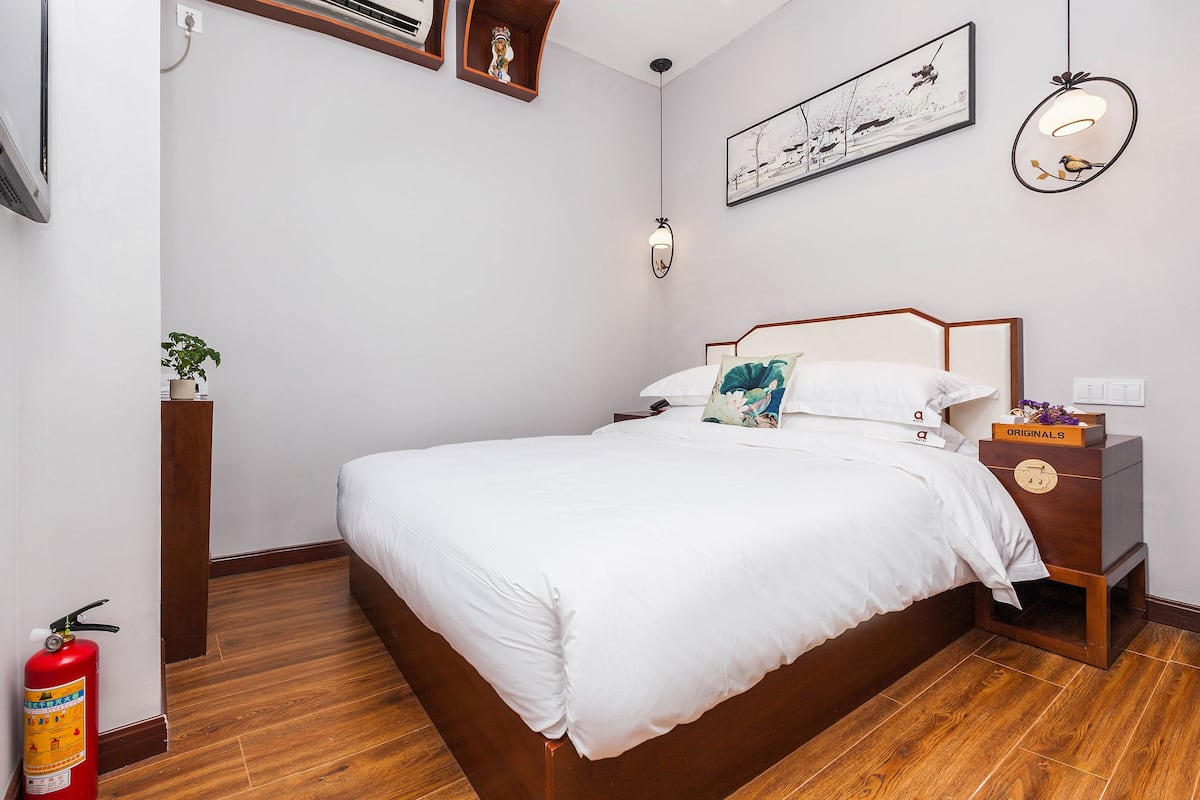 Une chambre à coucher bien rangée, avec un lit en bois, une table de chevet avec une lampe, une œuvre d’art encadrée au mur et une petite plante posée sur le sol.