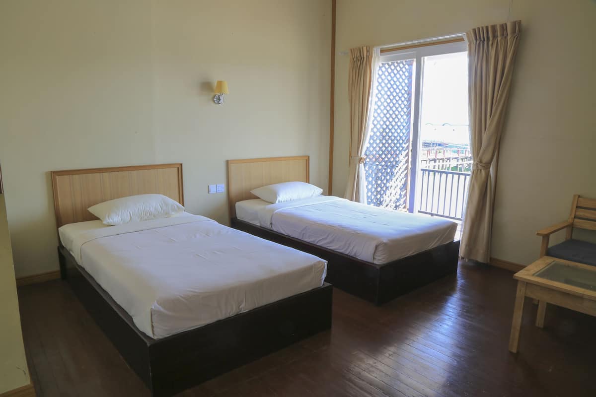 A hotel room with two single beds, a wooden floor, a small table, and a balcony with a view.