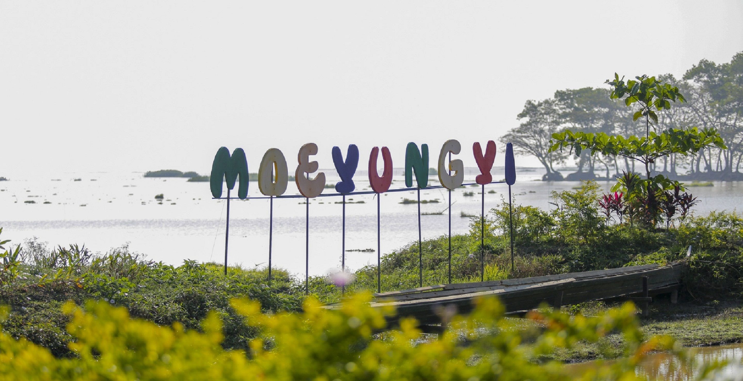 A sign with the name "MOEYONGYI" in colorful letters, surrounded by greenery and a body of water.