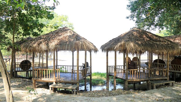 Two thatched-roof wooden structures with benches and railings, situated on a sandy beach with a water body in the background. - Moe Yun Gyi Wetlands Resort (Bago)