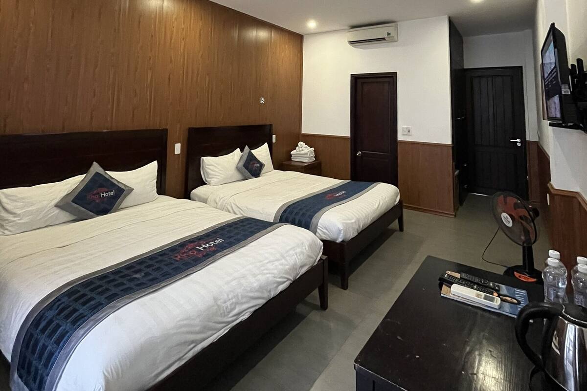 A hotel room with two beds, a small table with a kettle, and an air conditioning unit on the wall.