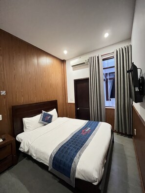 A hotel room with a wooden headboard, a bed with white bedding and a blue accent, an air conditioning unit, a TV mounted on the wall, and a window with curtains. - Kha Hotel - Hostel (Hue)