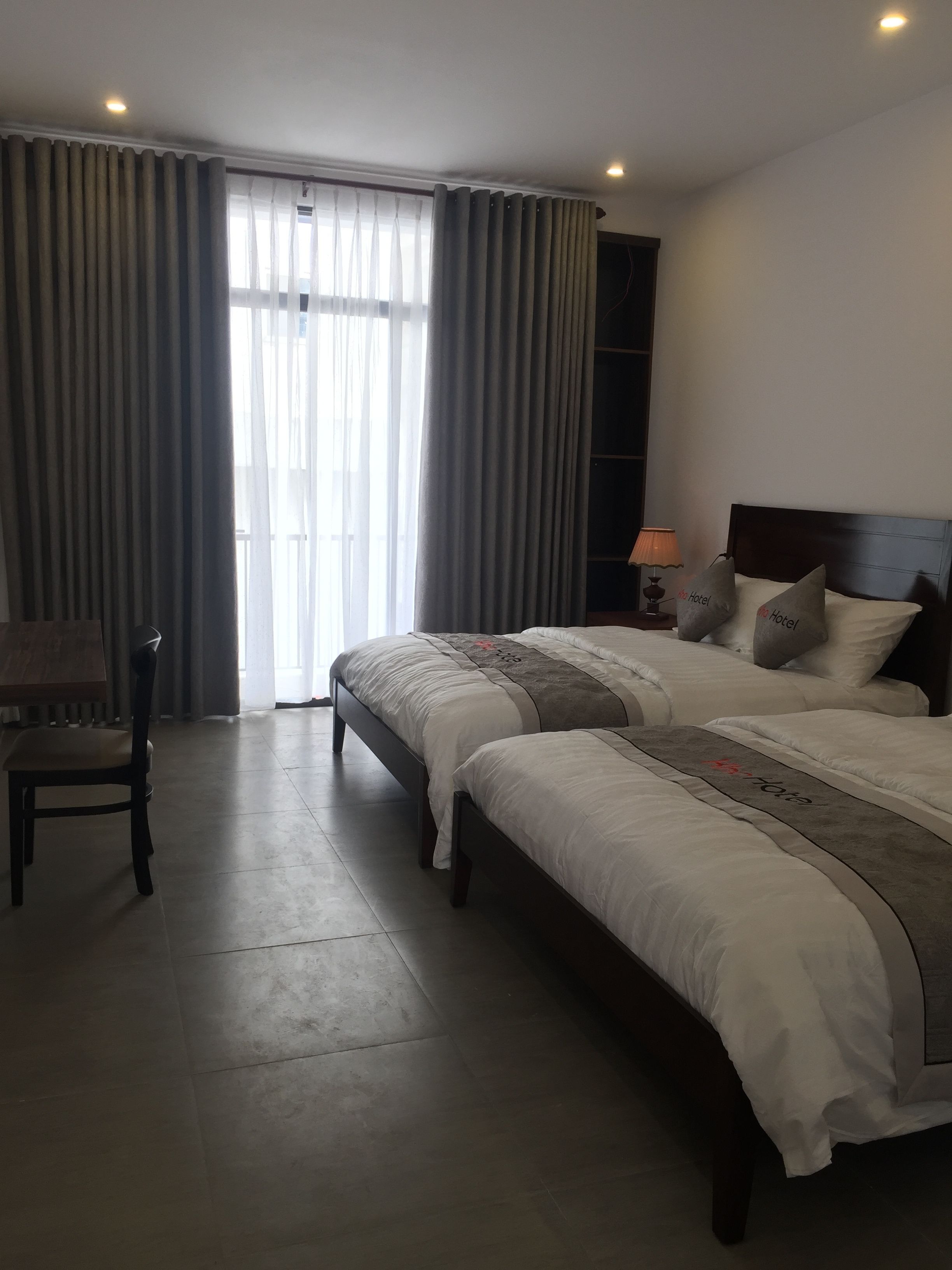 A hotel room with two beds, a chair, a nightstand with a lamp, and a large window with curtains.