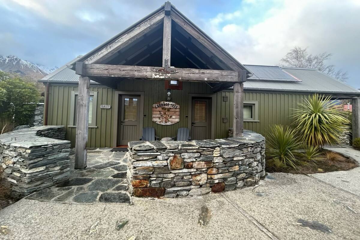Een rustieke hut met een stenen fundering, een houten veranda en een bordje met de tekst 'Cabbage Tree'.