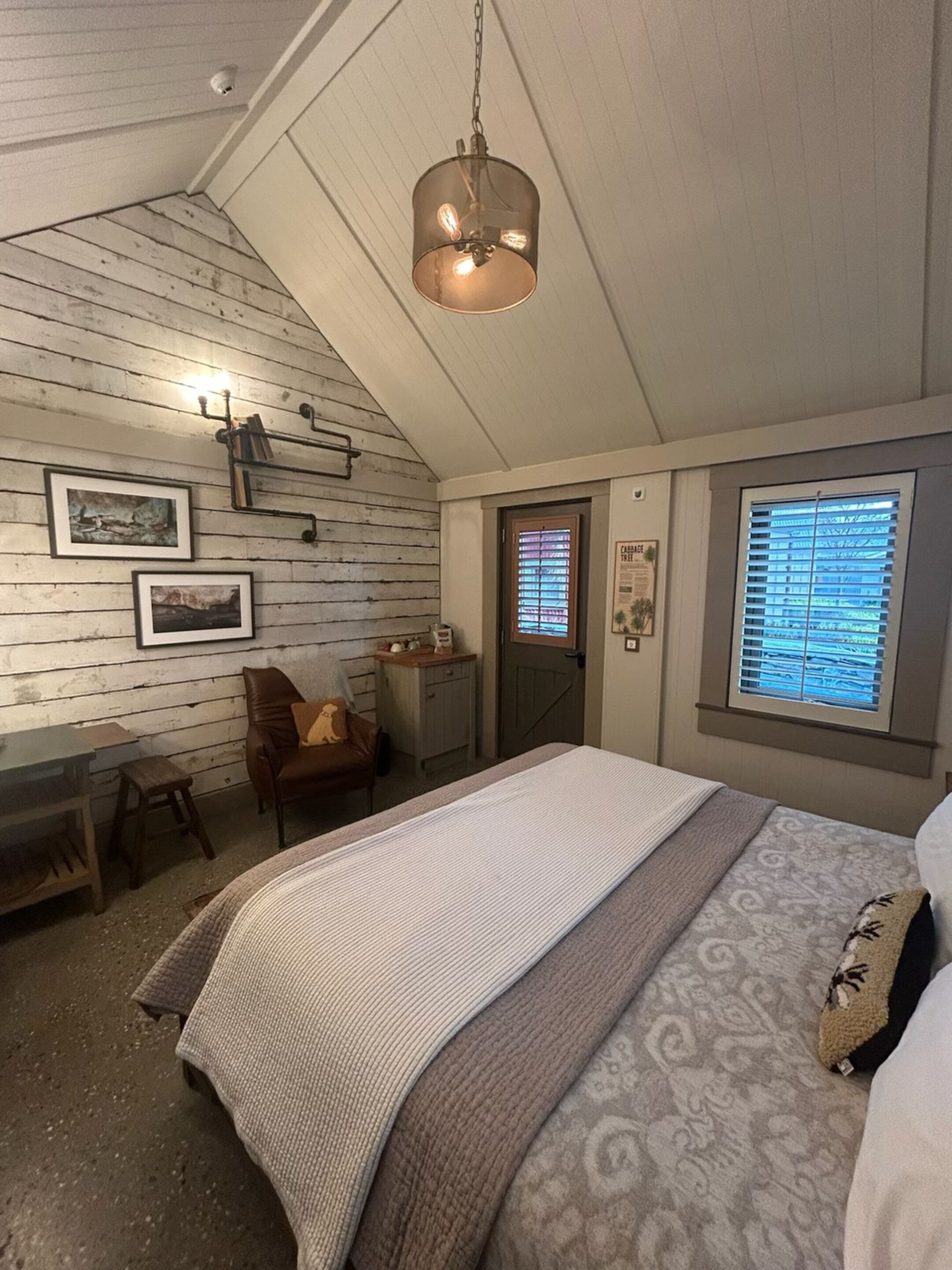 Une chambre confortable avec un plafond en pente, un lit recouvert d’une courtepointe à motifs, un bureau avec une chaise et une fenêtre avec des stores.