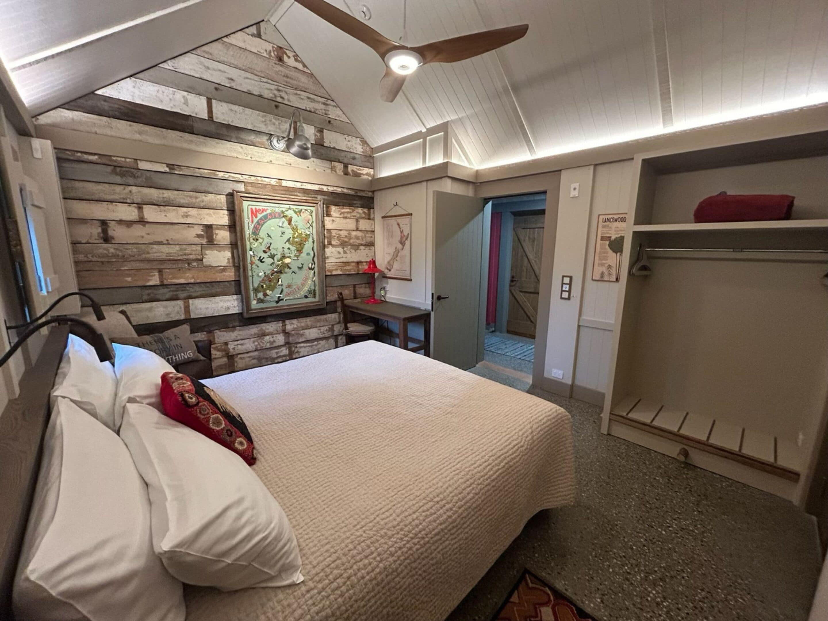 A bedroom with a bed, a wooden wall, a ceiling fan, a desk, and a door.
