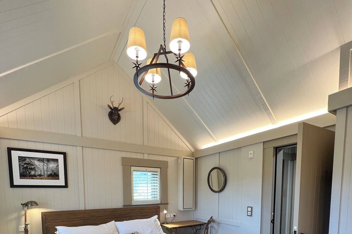 Een ruime slaapkamer met een groot bed, een houten hoofdeinde, een kroonluchter, een nachtkastje met een lamp, een spiegel en een deur naar een andere kamer.