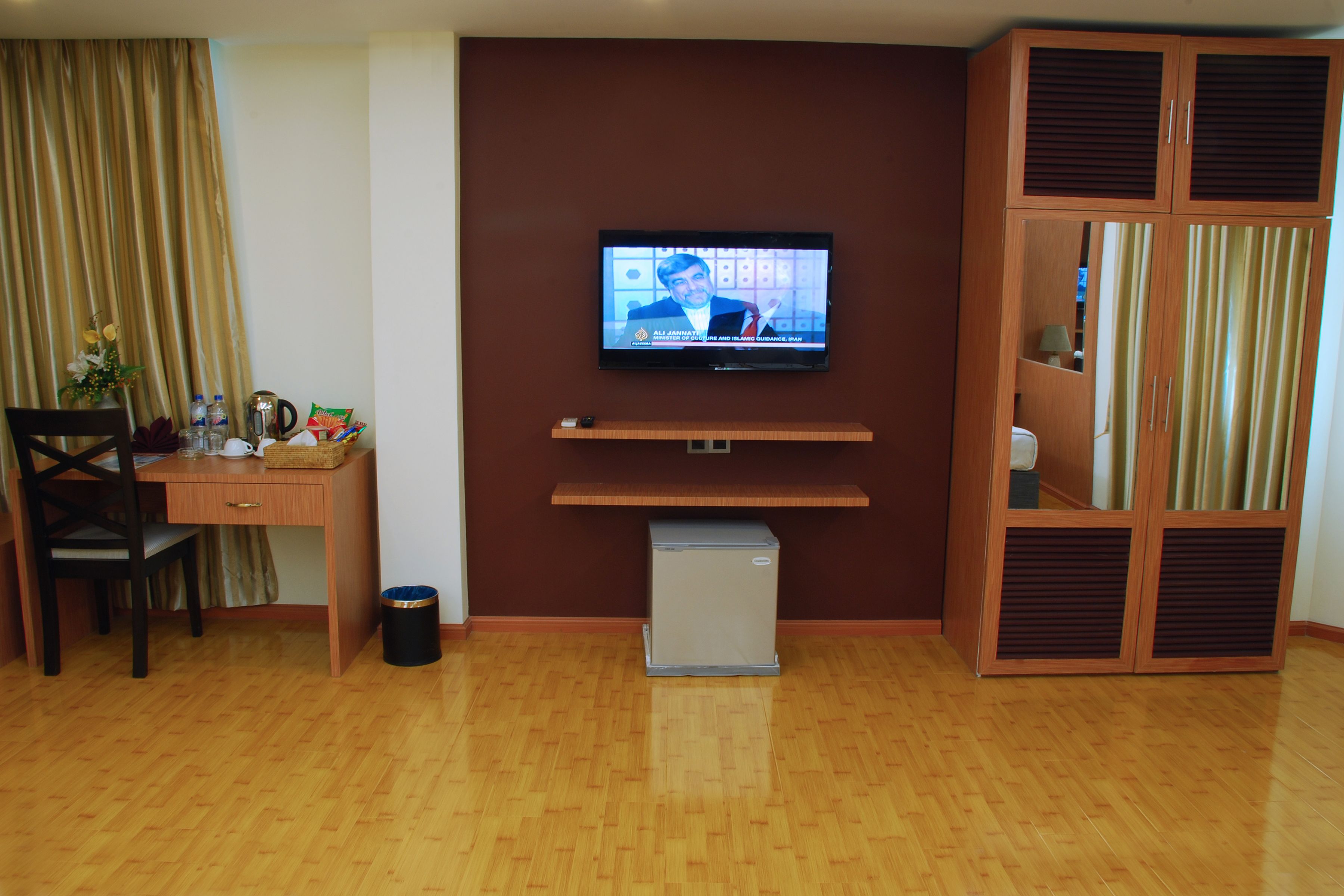 A living room with a television on the wall, a desk with a chair, a small trash bin, and a wardrobe.