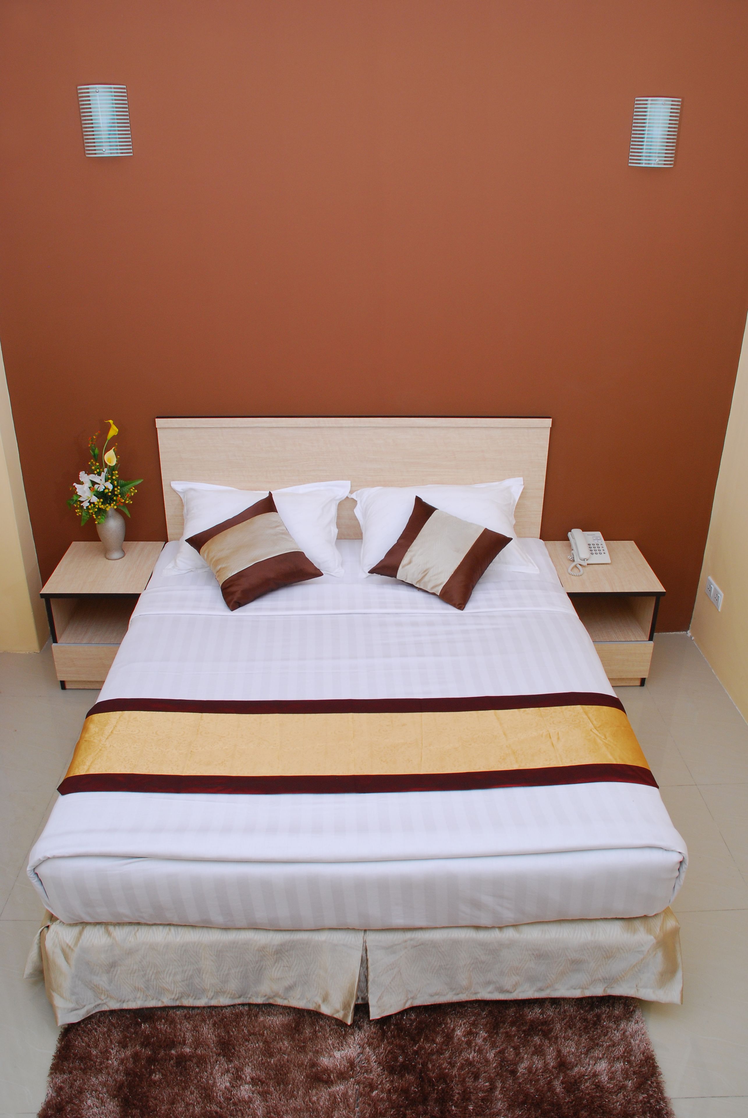 Una cama bien hecha con ropa de cama blanca y marrón, una mesita de noche de madera con un teléfono y un jarrón con flores.