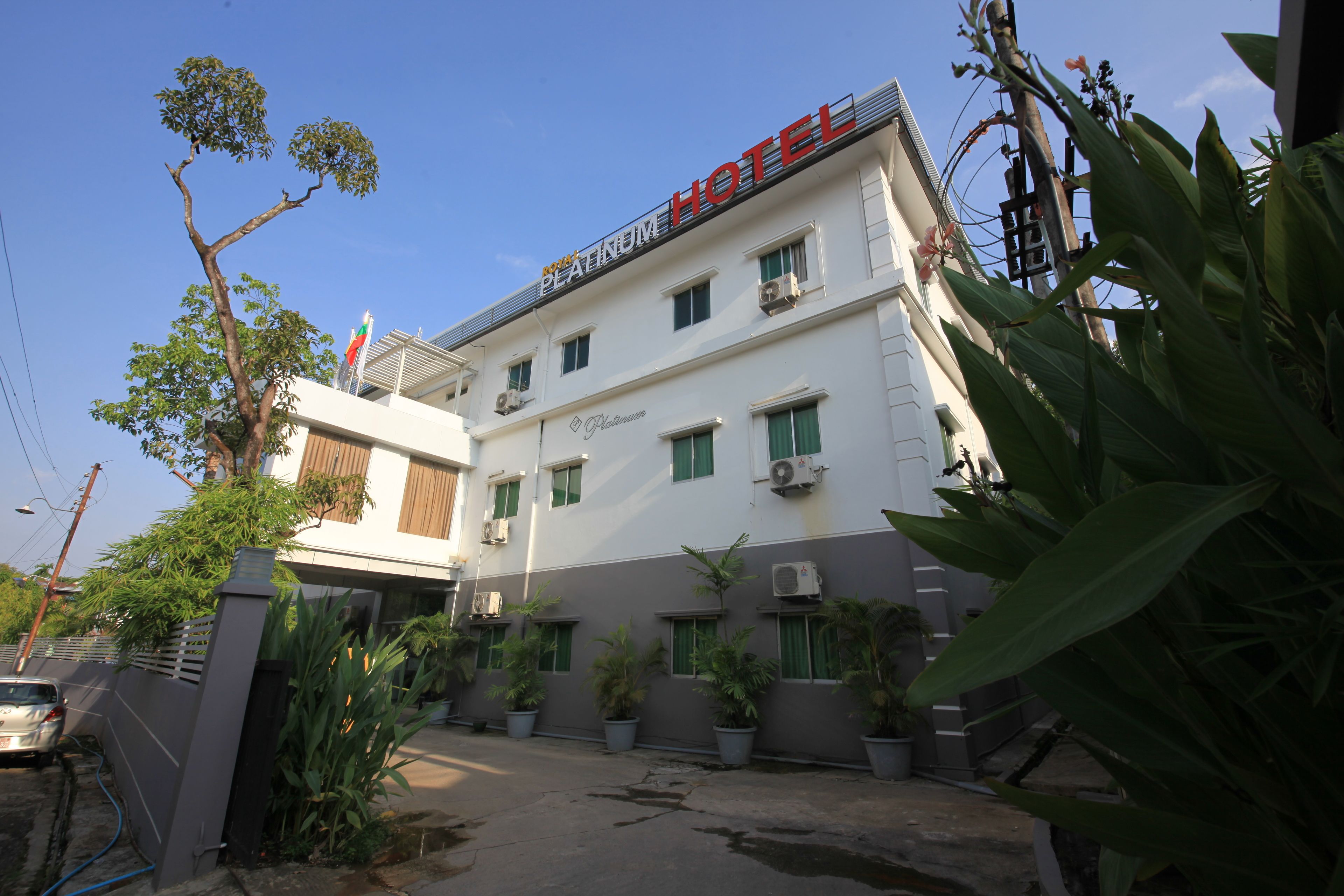 Un hotel con un letrero que dice 'PLATINUM HOTEL' y varios equipos de aire acondicionado. Hay plantas en macetas y un coche estacionado adelante.
