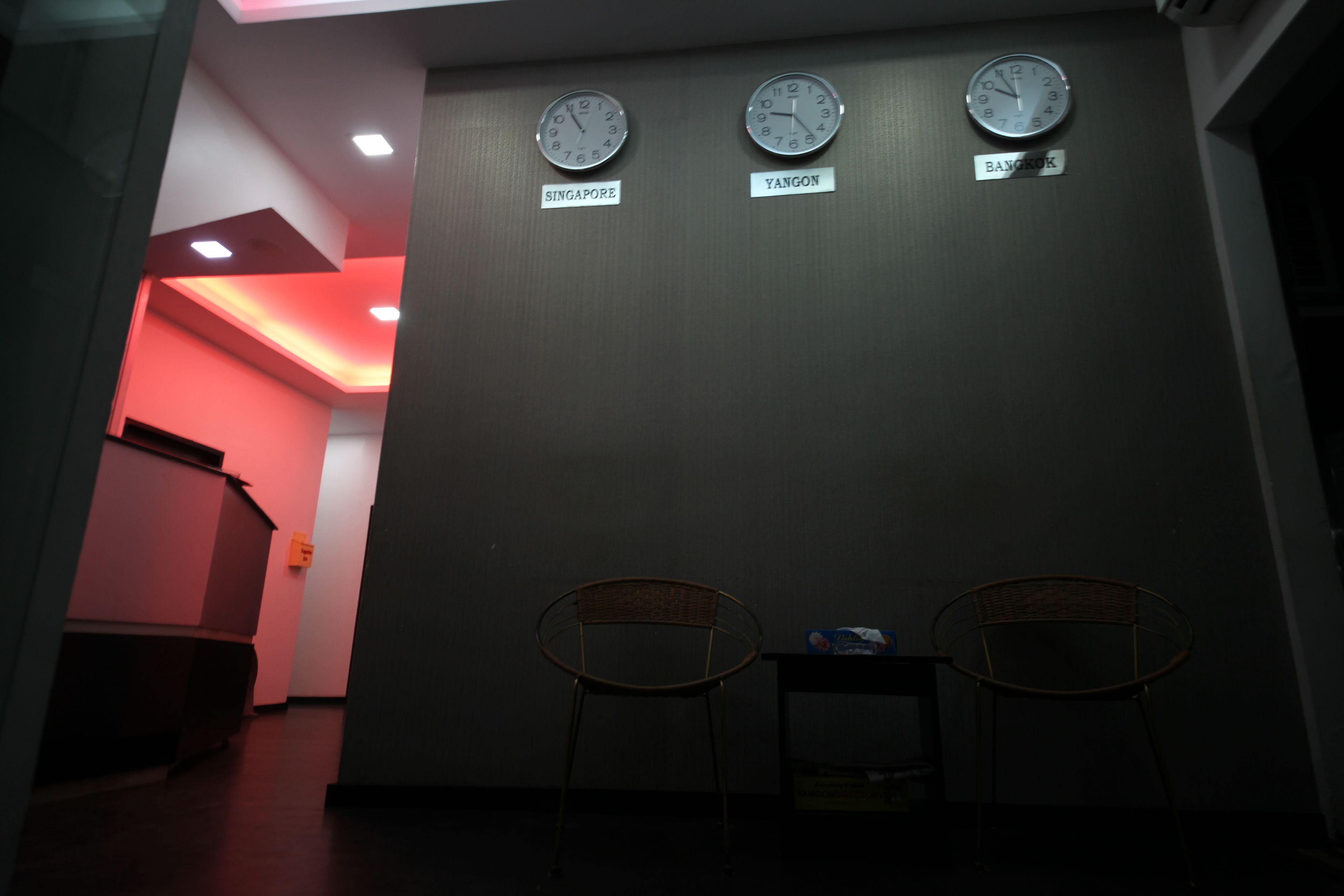 Un área de espera con dos sillas, tres relojes mostrando la hora de Singapur, Yangón y Bangkok, y un pasillo iluminado en rojo al fondo.