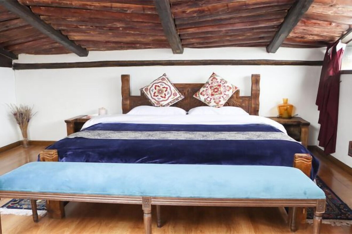 Quarto com cama de madeira, um banquinho, mesa de cabeceira e um objeto decorativo na parede.