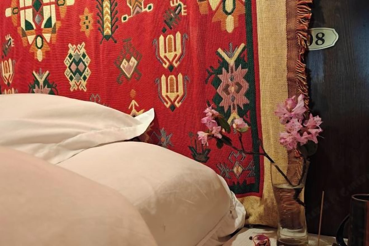 A bed with white linens, a red patterned curtain, a vase with pink flowers, and a door with the number 8.