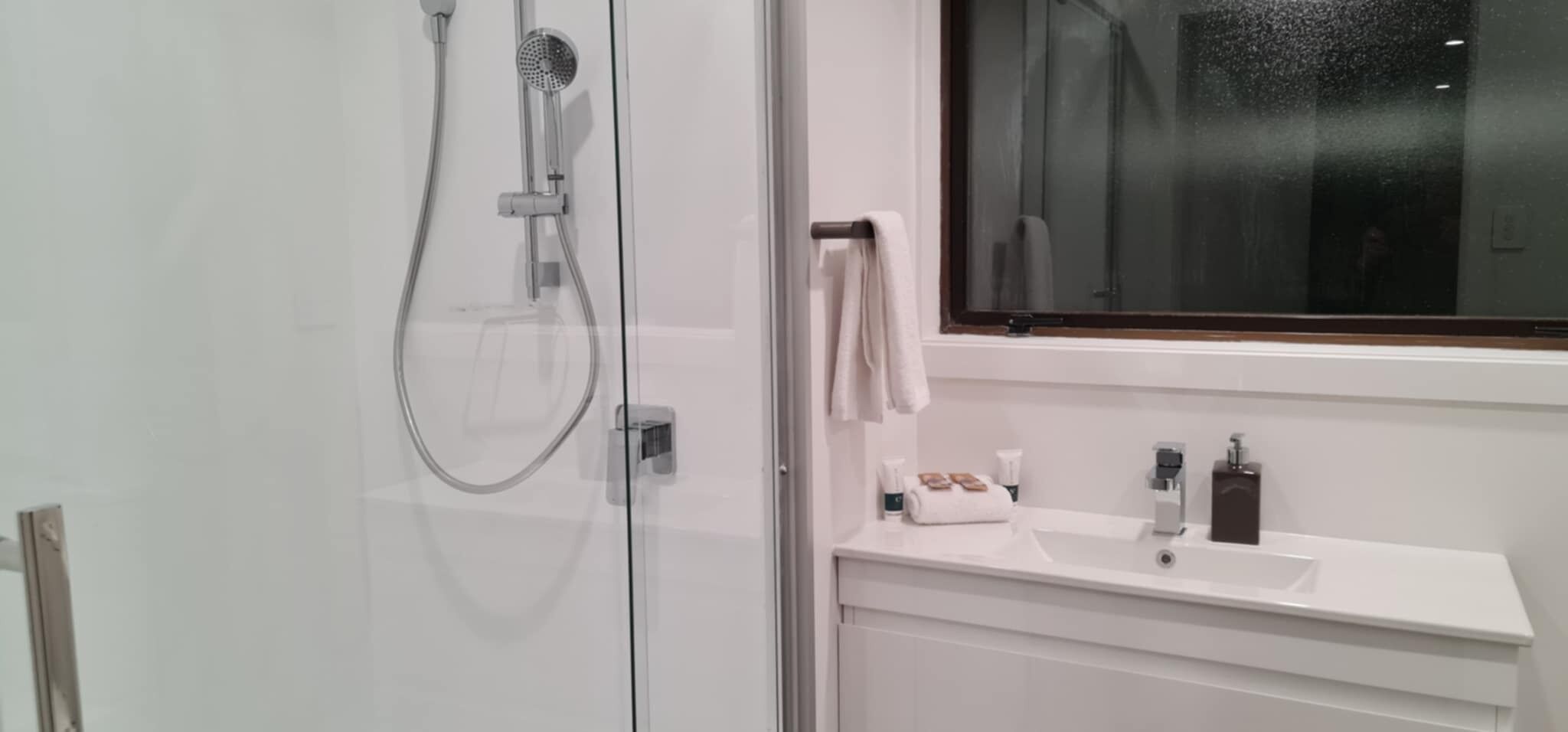 A modern bathroom with a glass shower enclosure, a white sink with toiletries, and a large window.