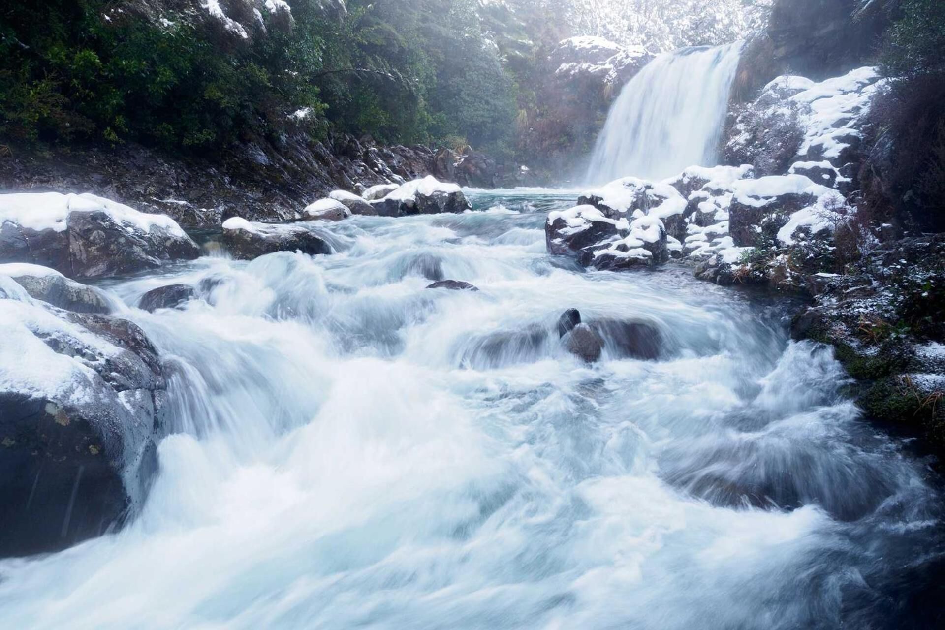A river with a waterfall surrounded by snow-covered rocks and greenery.