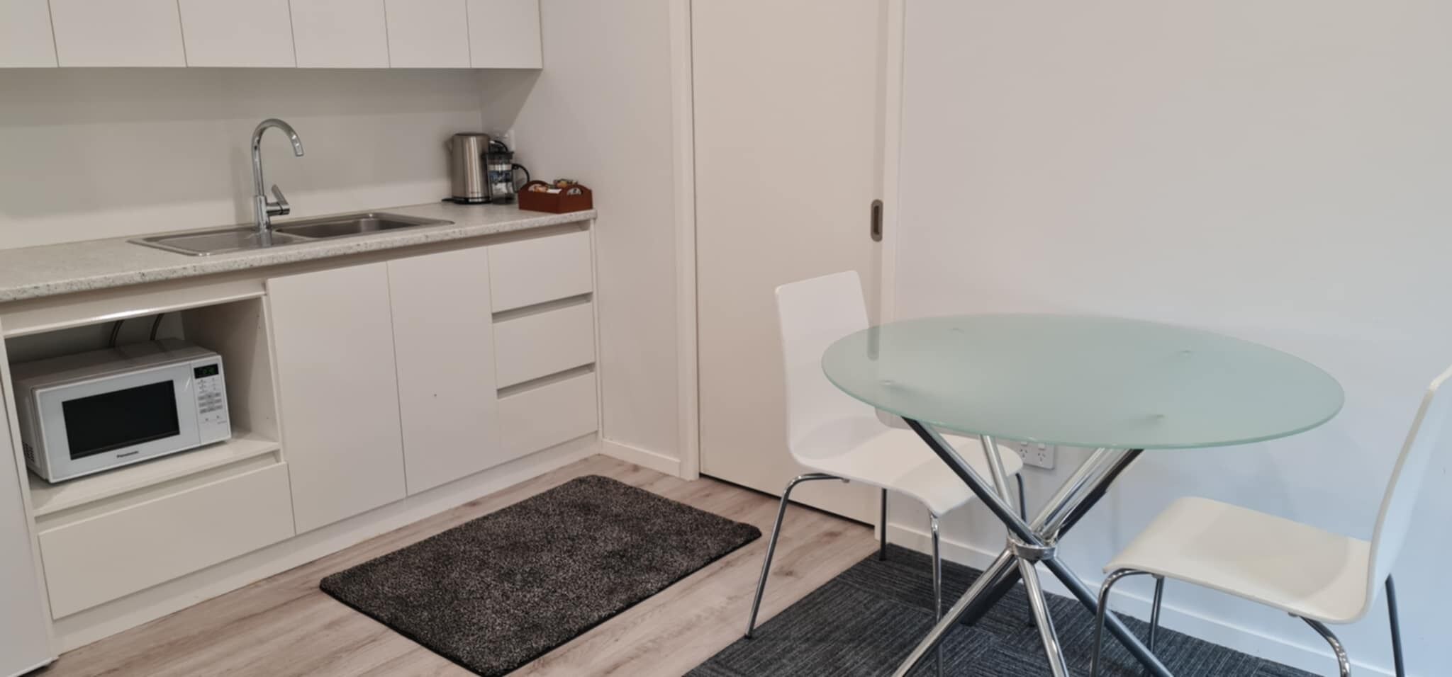 Cocina compacta con microondas, lavamanos y cafetera. Hay una mesa de vidrio con dos sillas y una alfombra oscura en el suelo.