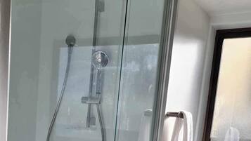 Une douche en verre avec un pommeau de douche à main et un robinet mural, un meuble-lavabo blanc avec un miroir et un plancher en bois.