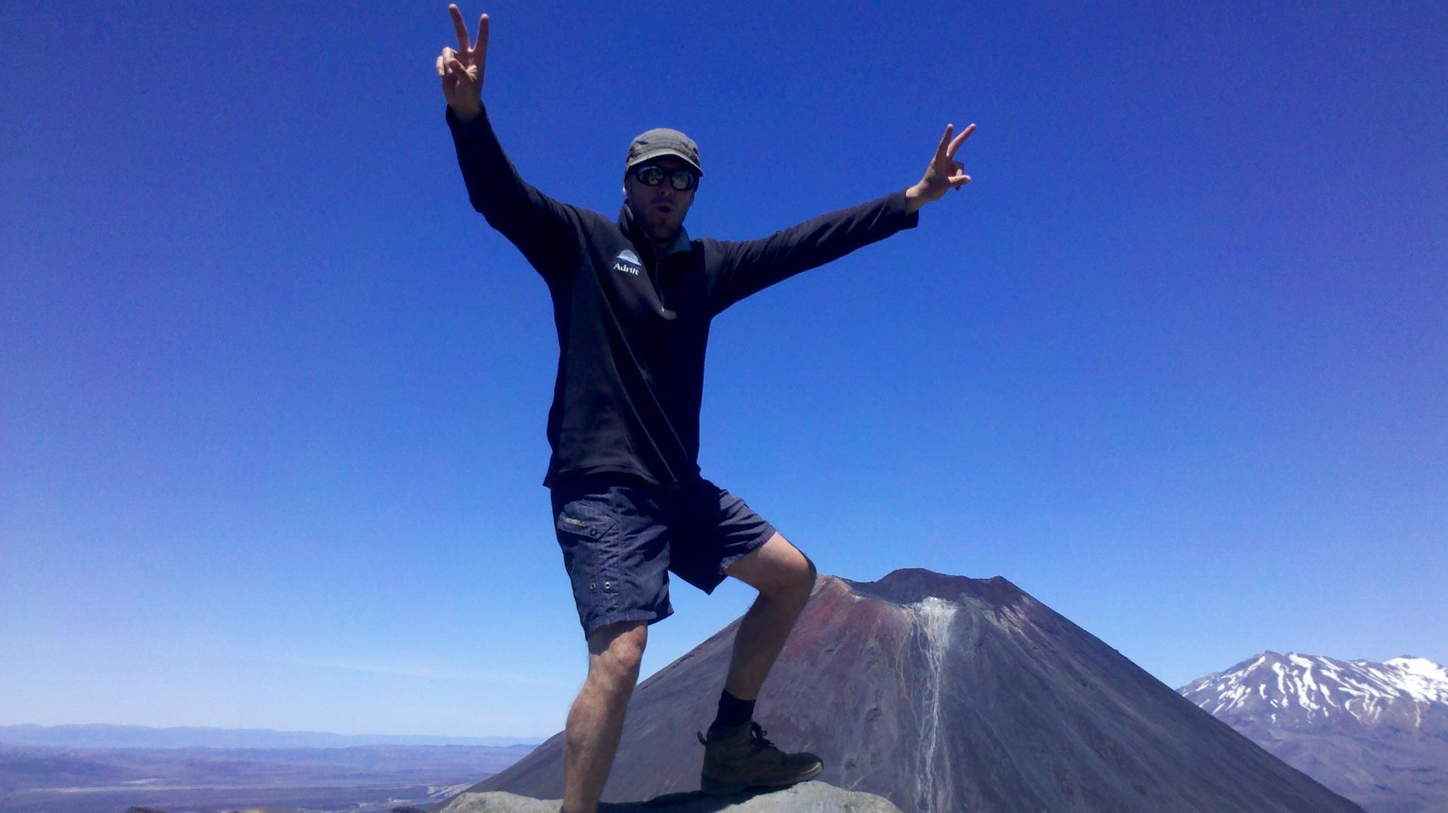 A person standing on a mountain peak with arms raised, wearing a cap, sunglasses, and a jacket.