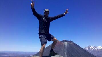 A person standing on a mountain peak with arms raised, wearing a cap, sunglasses, and a jacket.