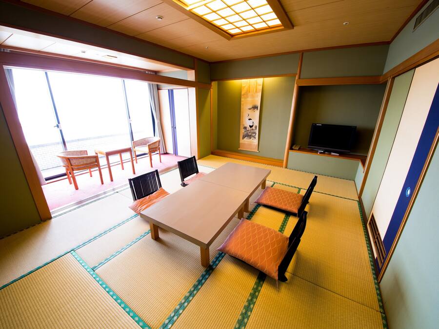 Una habitación tradicional japonesa con suelo de tatami, una mesa baja, sillas, un televisor y puertas correderas que dan a un balcón.
