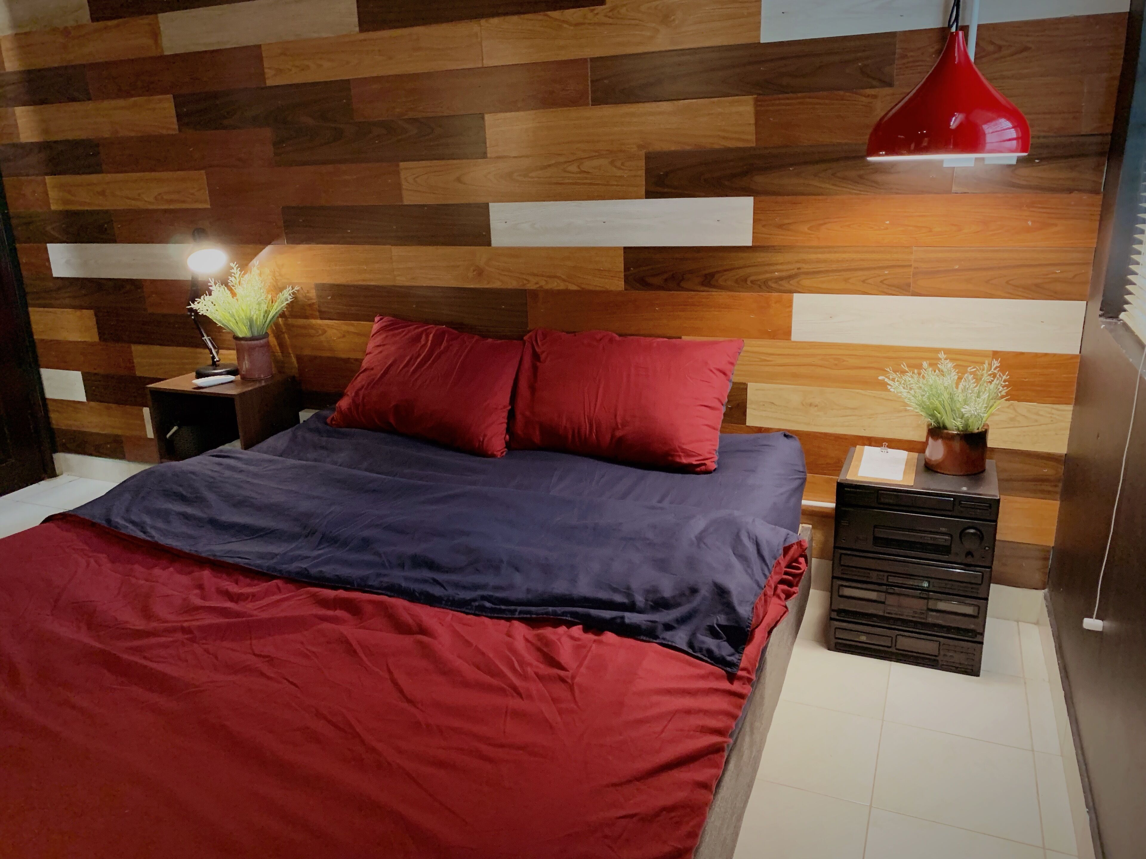 A modern bedroom with a wooden headboard, a red pendant light, a bedside table with a lamp, and a small potted plant.
