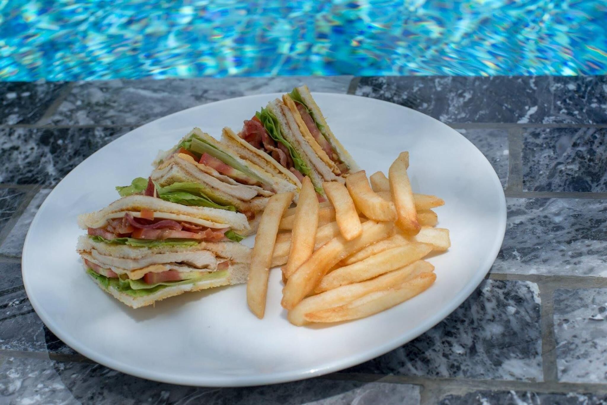 A plate with a sandwich and French fries by a poolside.
