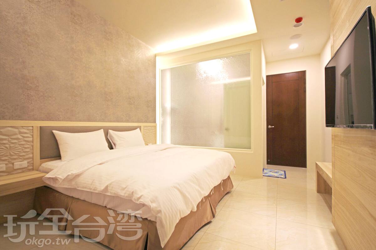 A bedroom with a large bed, a wooden nightstand, a TV, and a door leading to another room.