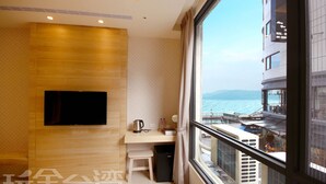Hotelkamer met een houten wand, een flatscreen televisie, een bureau met waterkoker en uitzicht op de zee door grote ramen.