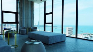 Habitación de hotel moderna con ventanas amplias, zona de estar con sofá y sillones, y vistas a la ciudad y al mar.