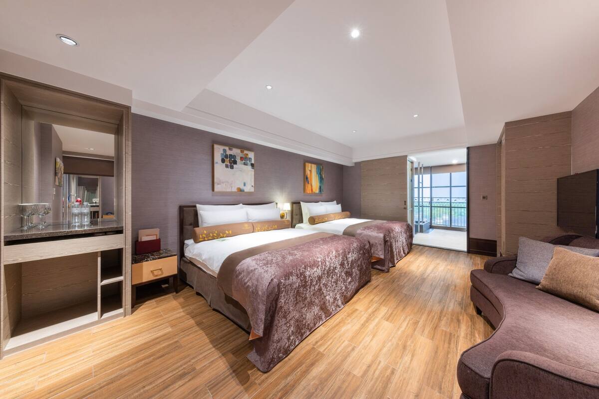 A modern hotel room with two beds, a desk, a sofa, and a large window.