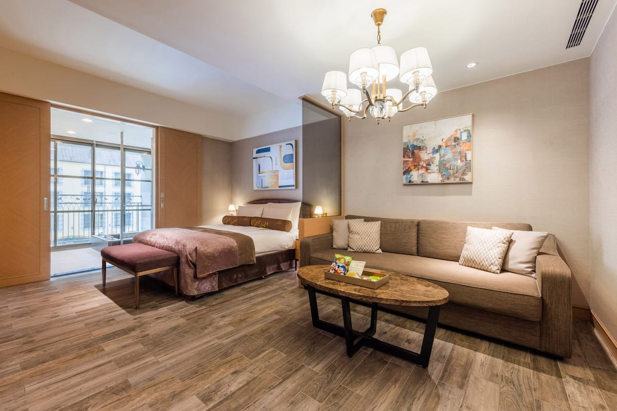 A modern hotel room with a bed, sofa, and a coffee table. There is a large window providing a view of the city.