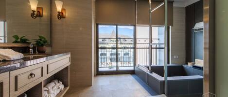 A modern bathroom with a large mirror, two wall-mounted lights, a bathtub, and a view of a building through a glass door.