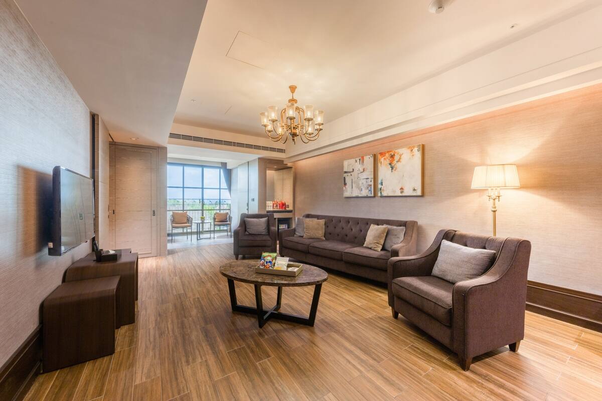 A modern living room with a sofa, coffee table, and a TV. There is a chandelier, artwork on the wall, and a lamp.