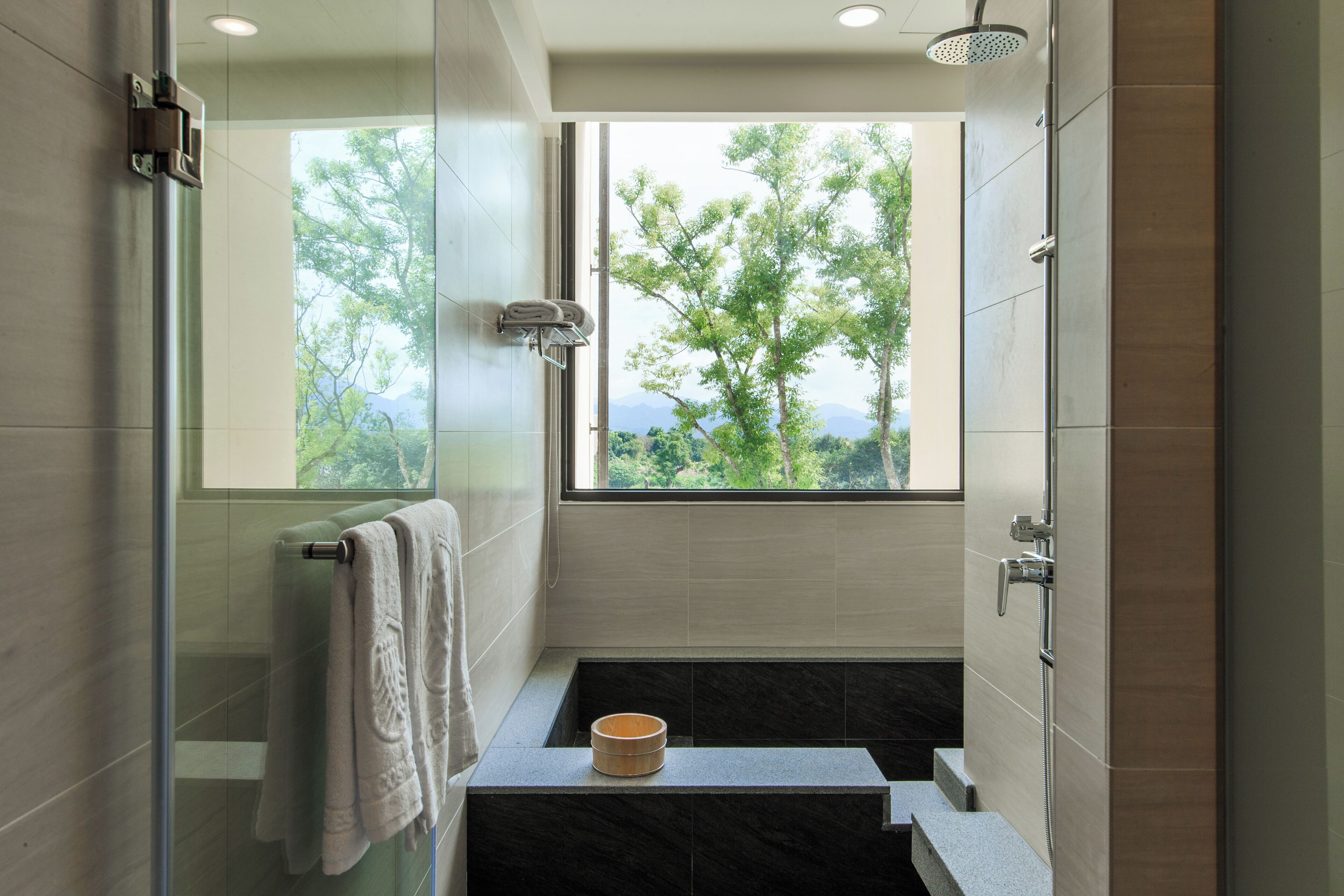 Un baño moderno con cabina de ducha de vidrio, una bañera con un cuenco de madera y un ventanal con vistas al verdor.