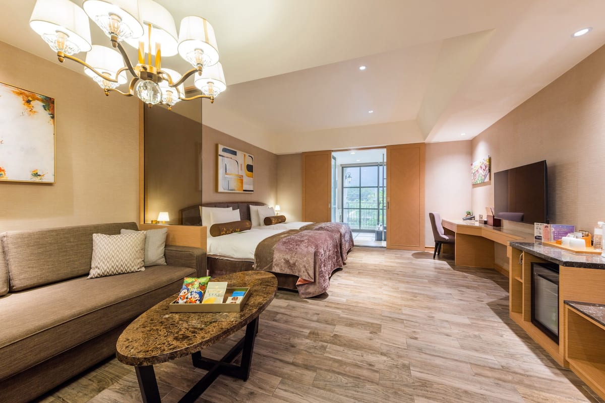 A modern hotel room with a sofa, a bed with a purple bedspread, a dining area with a TV, and a large window.