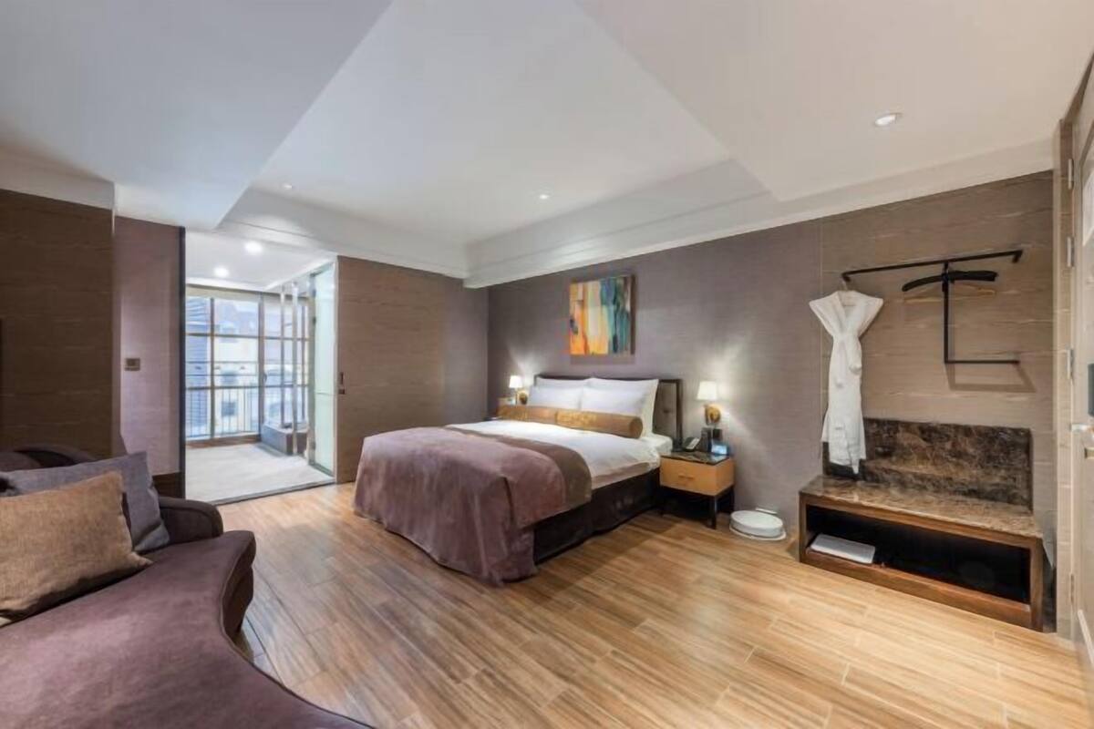 A modern hotel room with a large bed, a sofa, a bedside table, a robe, and a view of the city.