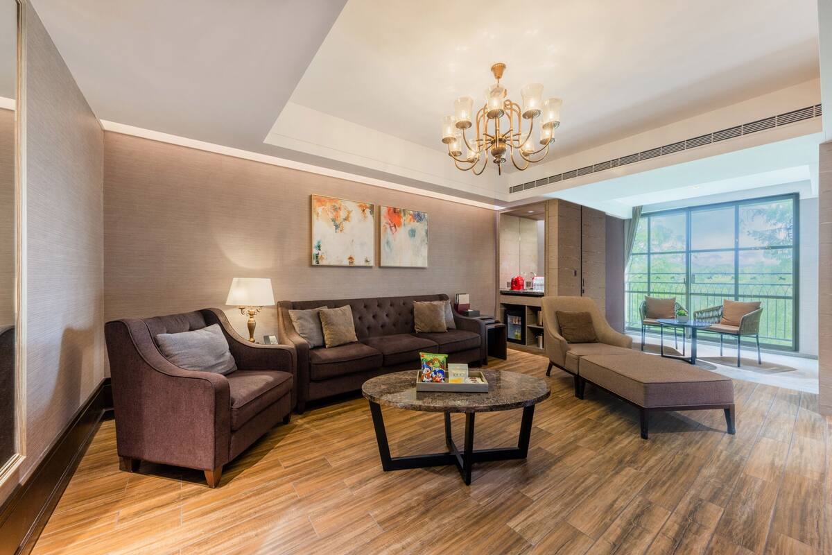 A modern living room with a sectional sofa, a chaise lounge, and a coffee table. There is a chandelier, artwork on the wall, and a large window providing a view.