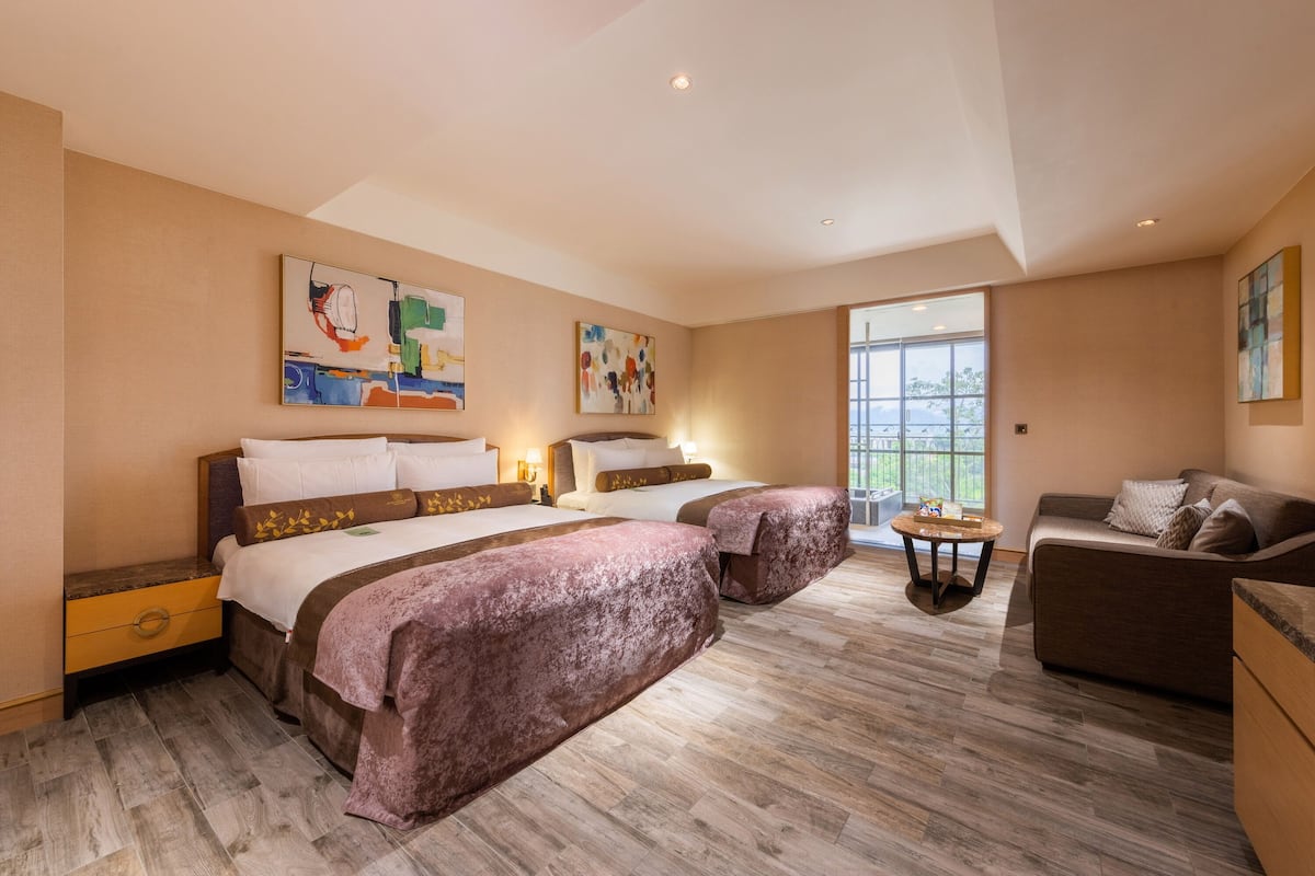 A hotel room with a large bed, a sofa, and two side tables. There are two paintings on the walls and a window providing natural light.