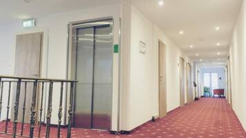 A hotel corridor with a red carpet, an elevator, and a staircase with a metal railing.