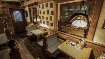 A rustic wooden interior with a dining area featuring wooden tables and chairs, a hanging light fixture, and a decorative wall with framed pictures.