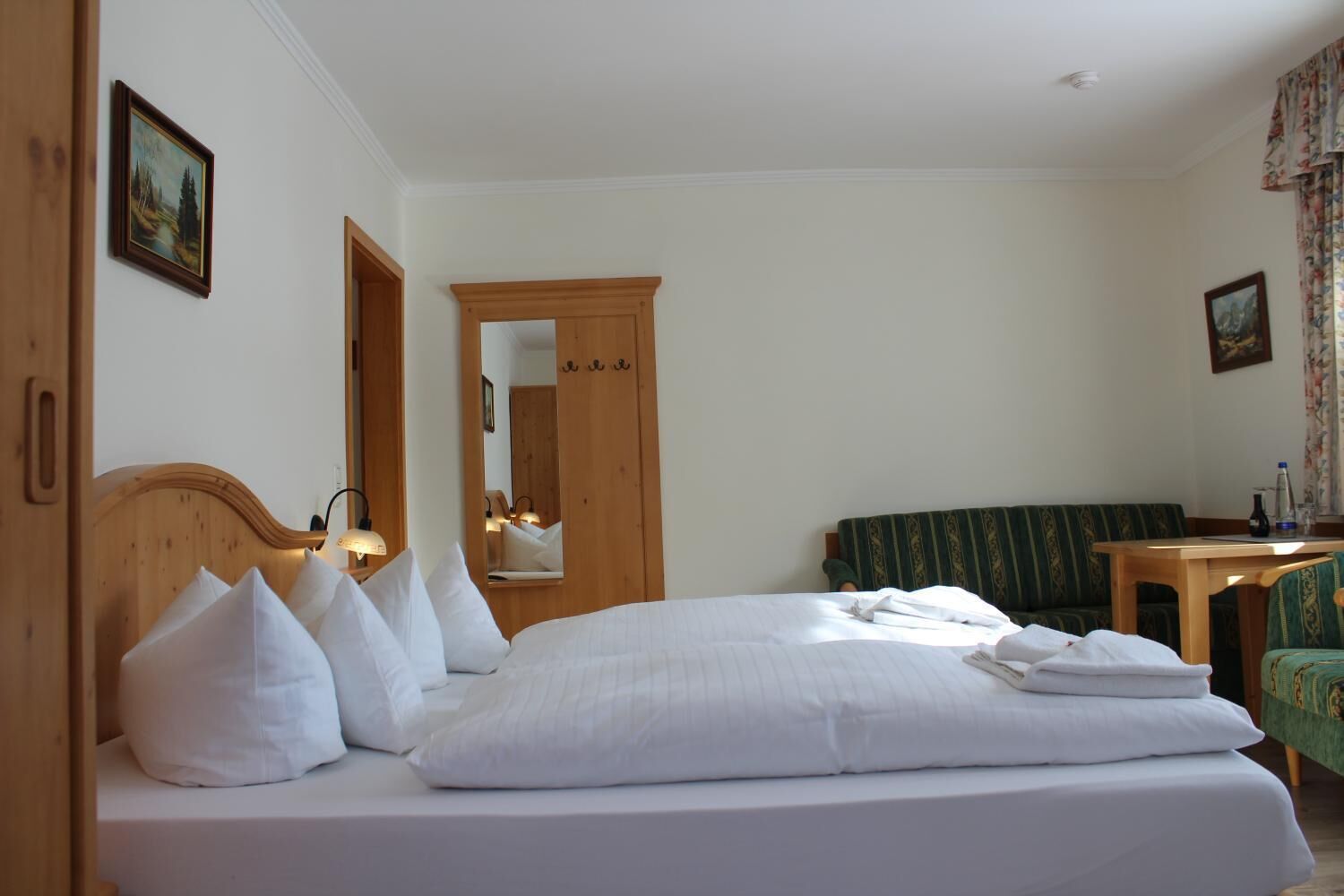 A hotel room with a large bed, a wooden headboard, a mirror, a green sofa, a small table, and a floral curtain.