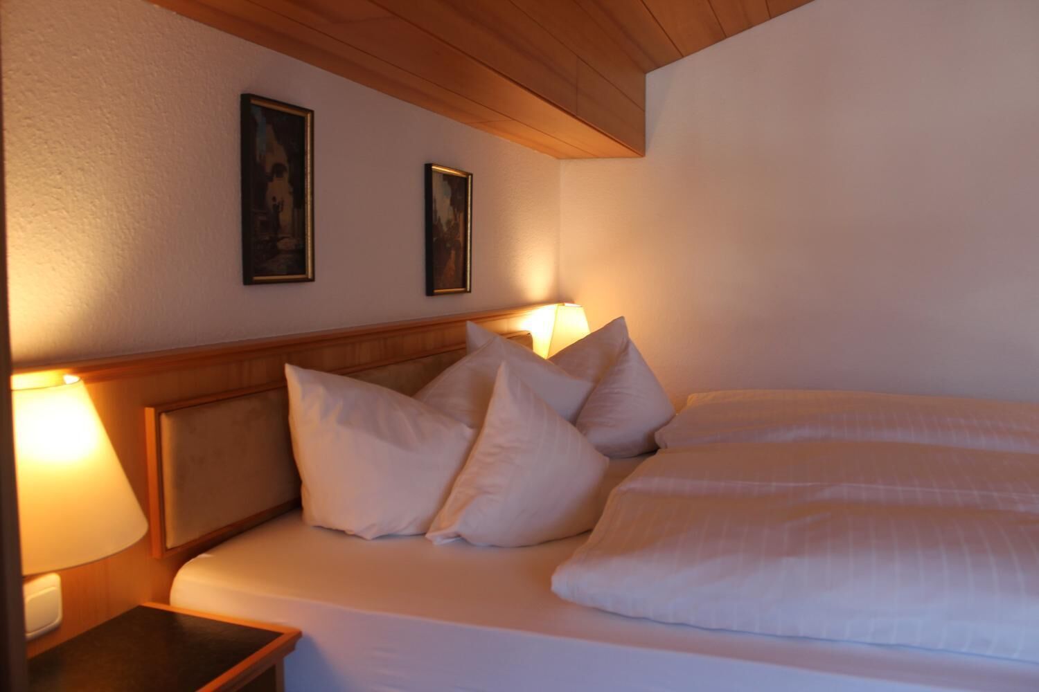 A hotel room with a wooden headboard, two bedside lamps, a bed with white linens, and two framed pictures on the wall.