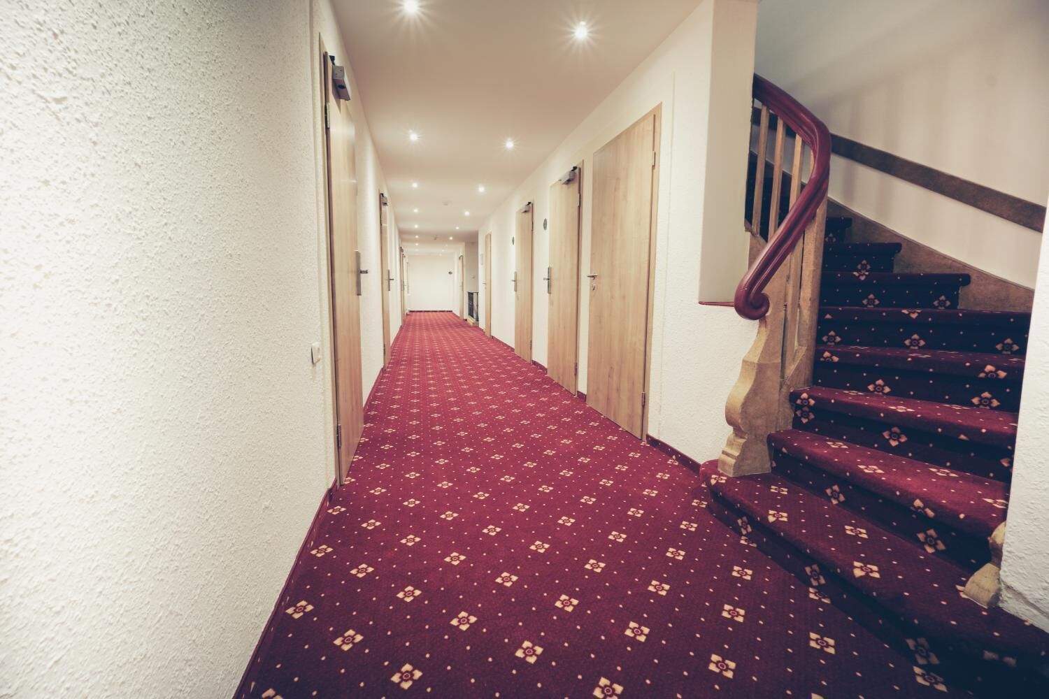 Ein Hotelkorridor mit rotem Teppich, Holztüren und einer Treppe mit einem hölzernen Geländer.