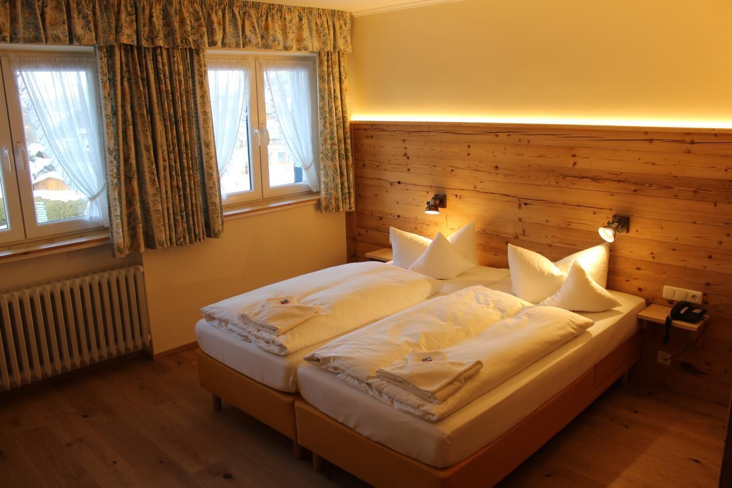 Uma cama de casal com lençóis brancos, cabeceira de madeira, abajures, janela com cortinas e um radiador.