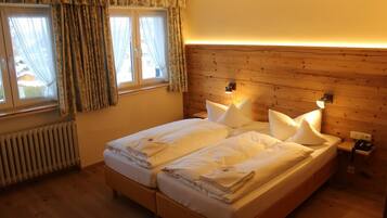 Uma cama de casal com lençóis brancos, cabeceira de madeira, abajures, janela com cortinas e um radiador.