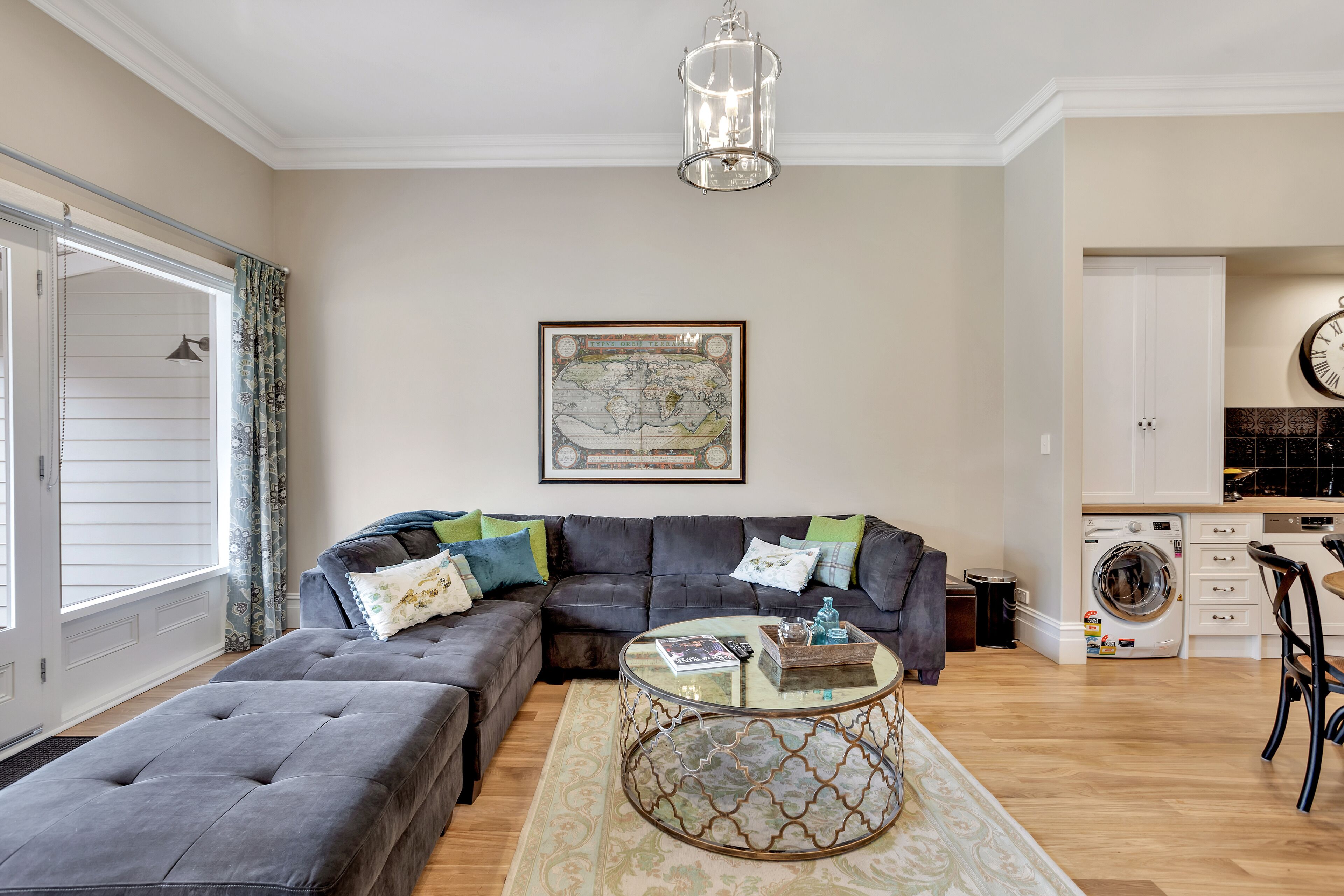 A living room with a sectional sofa, a glass coffee table, a map on the wall, a washing machine, and a dining area.